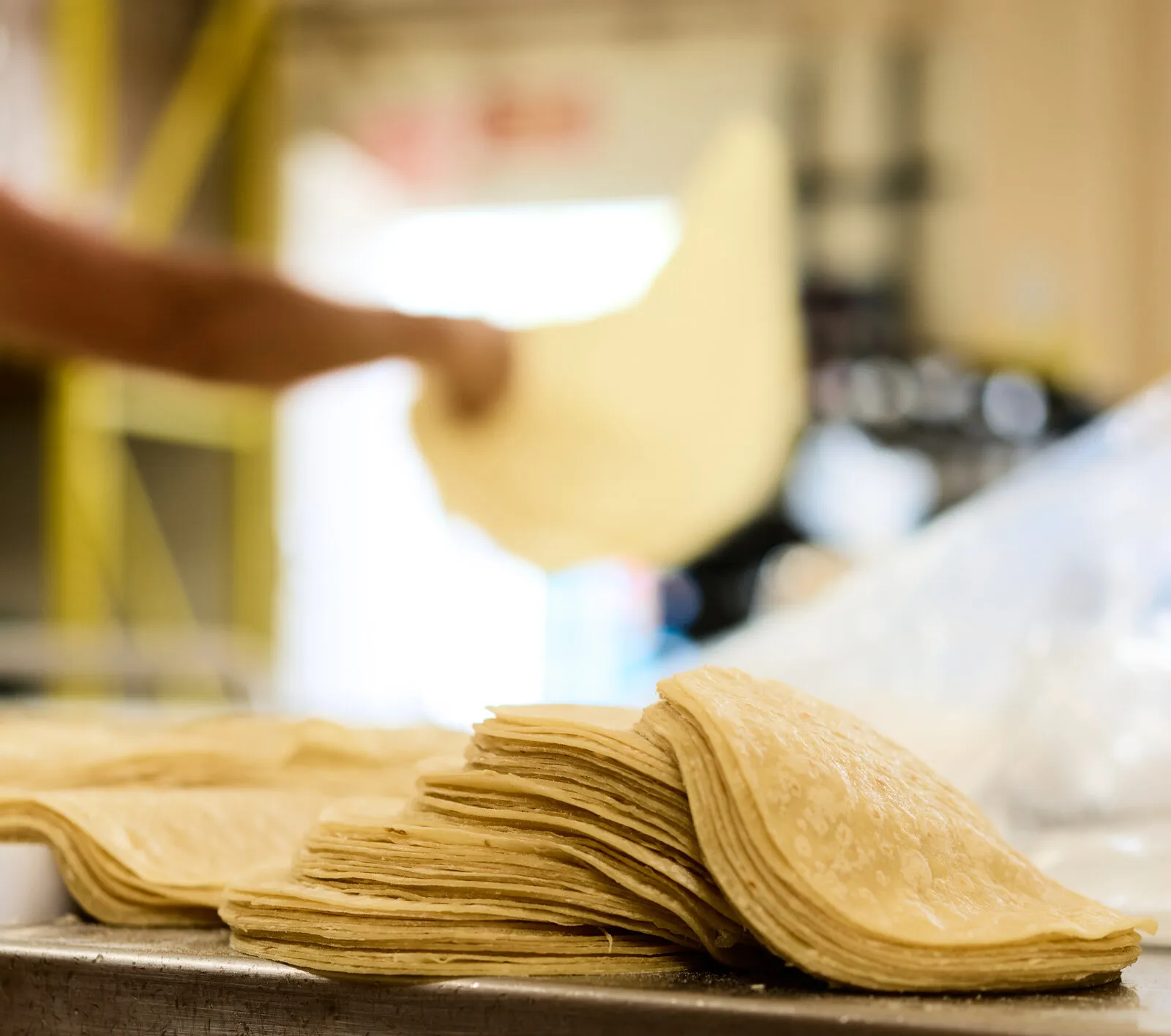 A stack of corn tortillas on a tray, with a hand lifting one behind it